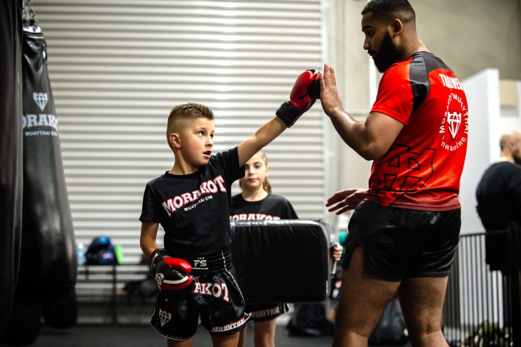 A young person wearing boxing gloves is practicing with an adult. Both are wearing the gym's logo shirts, with punching bags and other training equipment visible in the background. Morakot Muaythai Gym Epping Melbourne.