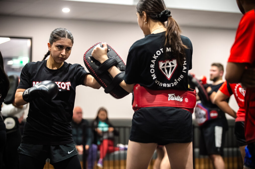 One person is throwing an elbow strike while the other holds a kick pad. Both are wearing black shirts with the gym's name and logo. Other participants and training equipment are visible in the background at Morakot Muaythai Gym Epping Melbourne.